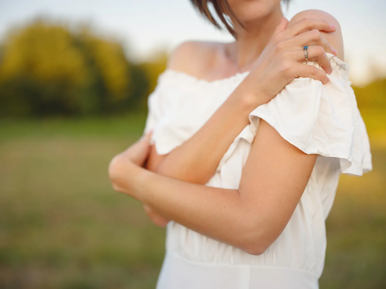 A woman resting one hand on her own shoulder, standing outdoors in warm evening light