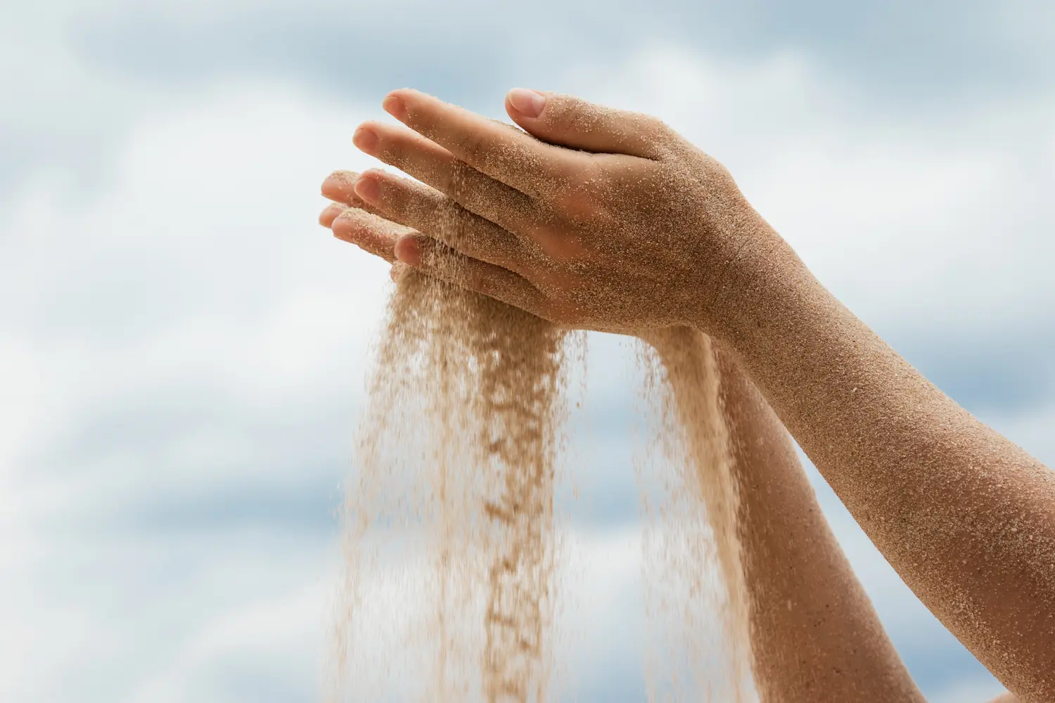 Hands letting fine sand fall through open fingers against a pale sky